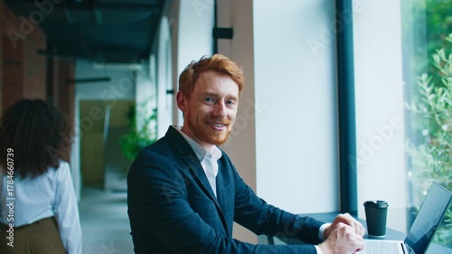 Attractive Caucasian businessman sitting near window using laptop and smiling confidently. Wearing formal suit. Bright modern corridor with plants and blurred colleague walking behind.