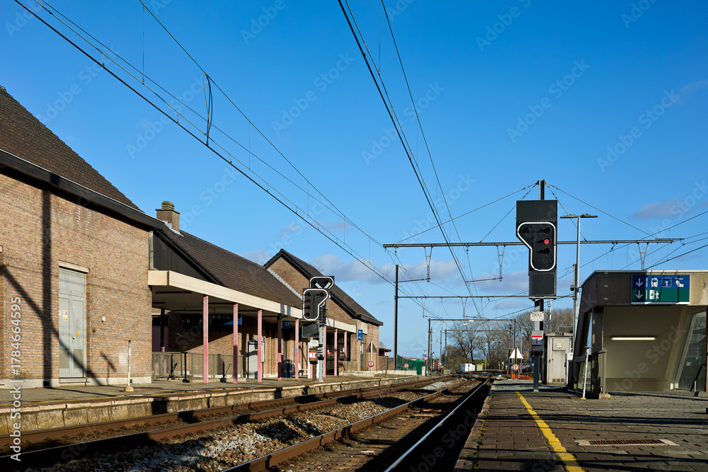 Fototapeta premium Bright, sunny day at a quiet train station with multiple tracks and platforms under a clear blue sky. Brick station buildings line the left, while modern elements and signal lights mark the right.