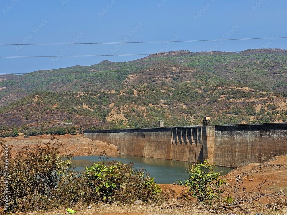 Obraz premium Mountain dam landscape Wide view of a dam surrounded by green hills and dry vegetation under bright sunlight, capturing natural engineering amidst rugged terrain.