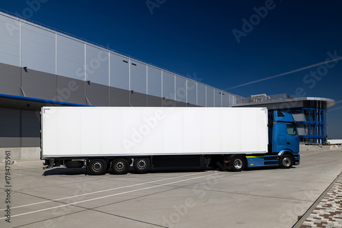 Blue truck with a white trailer parked in the yard of a logistics center. The truck is waiting to be loaded or unloaded into a warehouse. The photo was taken on a sunny day.