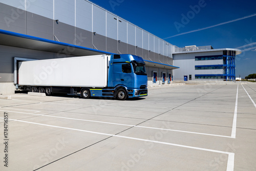 Blue truck with a white trailer parked in the yard of a logistics center. The truck is waiting to be loaded or unloaded into a warehouse. The photo was taken on a sunny day.