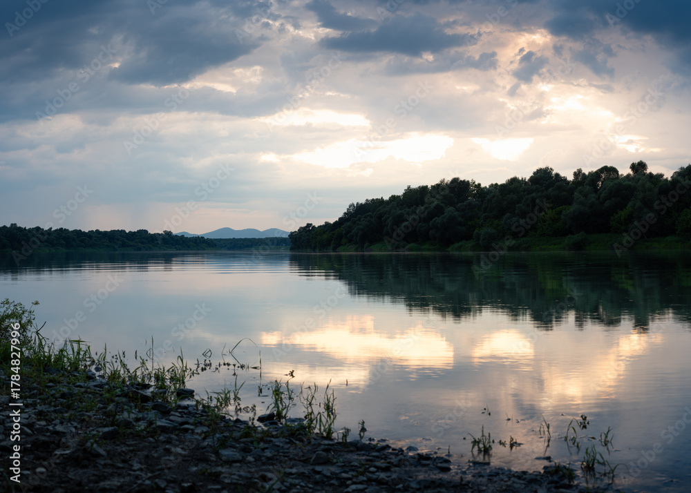 Fototapeta premium Landscape of Sava river and mountain in distance during sunset, glow in clouds reflecting in water surface