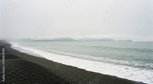 Fototapeta Naklejka Na Ścianę i Meble -  Black pebble beach on a foggy coastline. Gentle ocean waves with white sea foam. Misty mountains on the horizon. Minimalist nature landscape for wellness and tranquility concepts