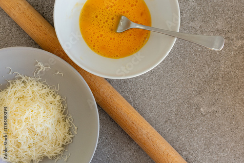 Grated cheese and beaten egg in bowls with rolling pin on gray counter, top view
