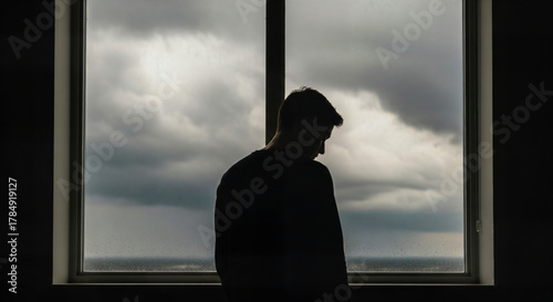 Pensive man standing alone gazing out window during stormy weather reflecting loneliness and introspection, emotional depth