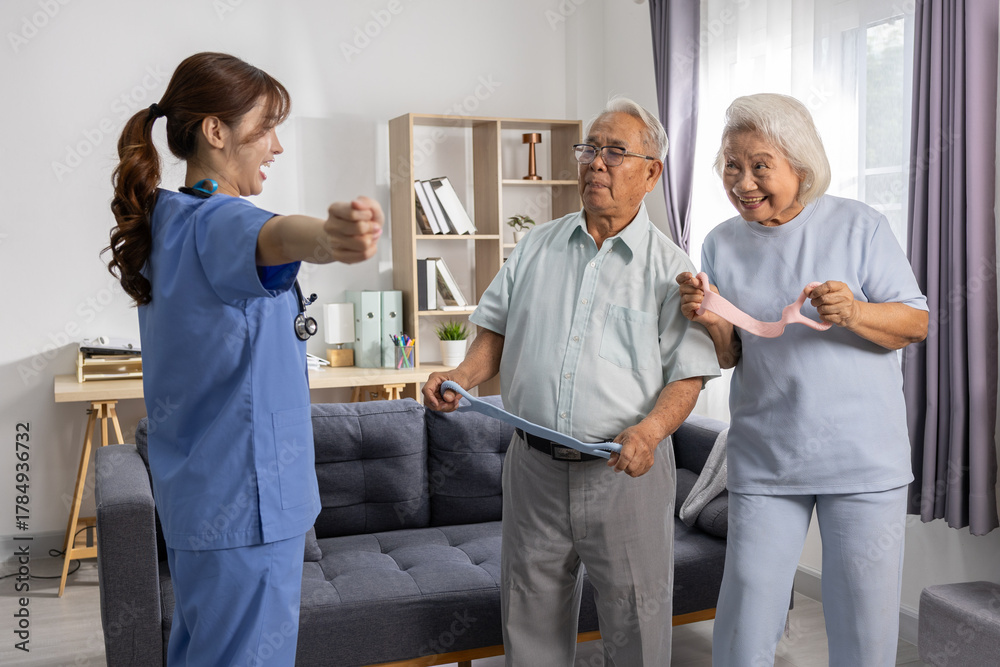 Obraz premium Asian Elderly couple doing resistance band exercises with female caregiver in cozy living room, smiling and following guided physical therapy routine with light equipment and natural light