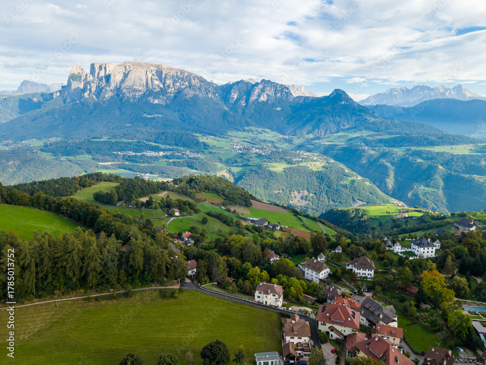 Fototapeta premium Aerial view of a scenic alpine village surrounded by green hills and mountains under a bright summer sky, South Tyrol, Italy