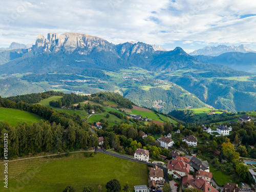 Aerial view of a scenic alpine village surrounded by green hills and mountains under a bright summer sky, South Tyrol, Italy