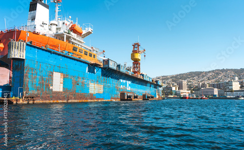 Large dry dock with crane and rusting ship hull in shipyard, industrial construction and maritime repair services, vibrant blue sky and coastal city view.