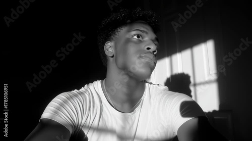 Young adult man in white tee looks up in dark hall with window shadows; indoor, all-season moody scene.