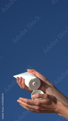 Female hands holding white mockup tube, opening it and applying cream on palm of their hand against clear blue sky. Concept of daily face cream for natural beauty, hydration, and lifestyle skincare