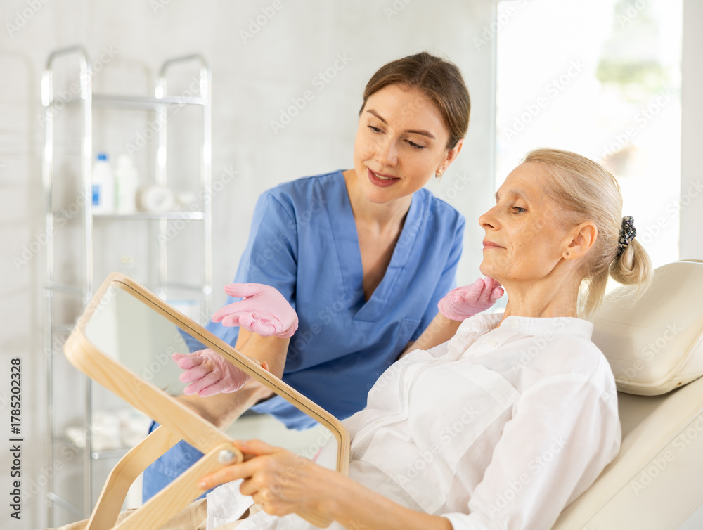 Obraz premium Elderly female patient sitting on examination couch in medical aesthetic office looking at mirror while professional woman cosmetologist explaining future hardware rejuvenating facial procedure