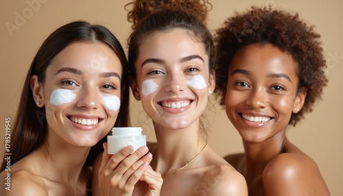 Three smiling diverse women apply white face cream in studio. Skincare routine for healthy glowing skin. Beauty products for pampering and self care.