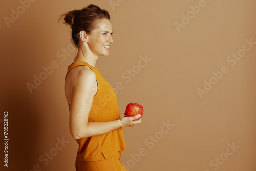 A fair-skinned woman in bright orange activewear smiles, holding a red apple in profile against a soft brown backdrop. The image conveys health, wellness, and self-care.