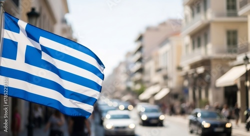 Fototapeta Naklejka Na Ścianę i Meble -  Greek flag waving with city street and car traffic in blurred background. National symbol for independence day, travel, and tourism.