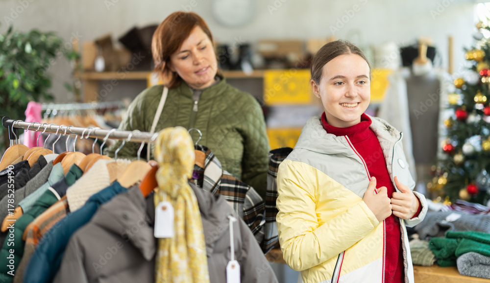Obraz premium Teen girl try on down coat in front of mirror in clothes store, shopping with mother, X mas sale. Customer is trying on clothes in store. Buyer is immersed in choice of clothes. .