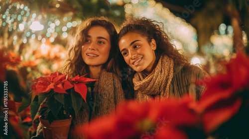 Two Young Women Smiling and Admiring the Vivid Red Poinsettias, or Flor de Nochebuena, at a Festive Christmas Market in Mexico, Surrounded by Sparkling Lights and Holiday Cheer
