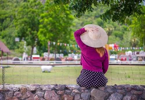 Young woman wearing straw hat sitting on rock wall looking at scenic park view. Rear view of a female tourist relaxing outdoors, enjoying the beautiful view of farm or theme park on sunny vacation day