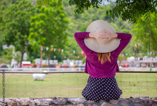 Woman in straw hat relaxing with hands behind head enjoying peaceful park view. Young female tourist sitting comfortably on rock wall, hands clasped behind head, looking at tranquil outdoor landscape.