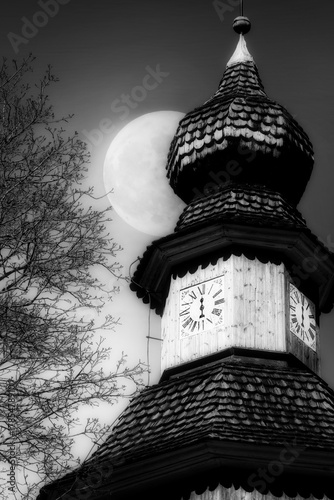 Church bell tower with old mechanical clock with only an hour hand at Midnight, with Full Moon on the sky. Monochrome, vertical view.