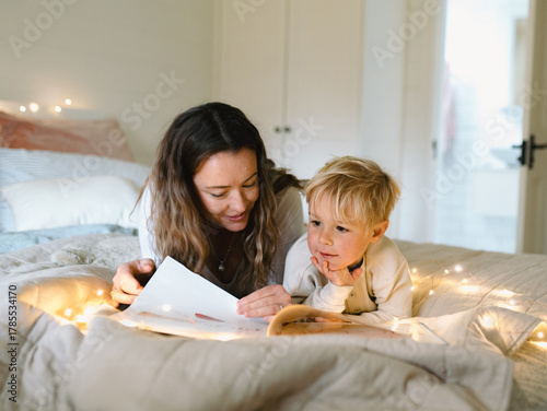 Mum and son lying on their belly on the bed with string lights reading a book