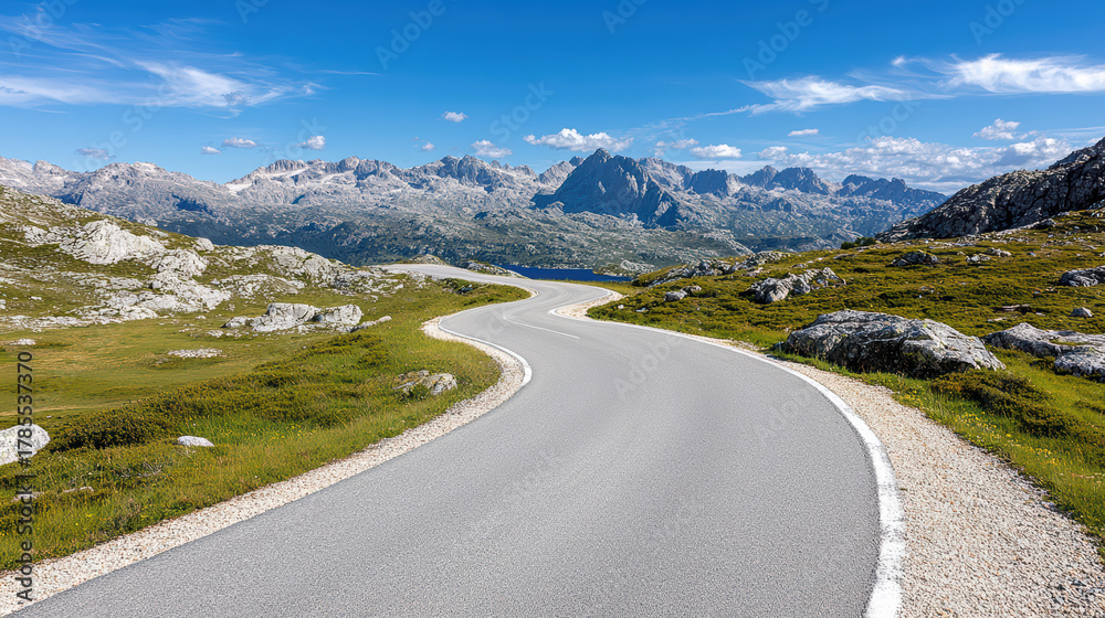 Fototapeta premium Winding mountain road with sharp turns offers panoramic view of rugged peaks under clear blue sky, surrounded by rocky terrain and green grass