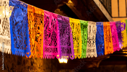 A colorful string of paper lanterns hang from the ceiling. The lanterns are of different colors. Mexican Day of the Dead tradition, offerings, food, colors, decorations, and papel picado, La Catrina.