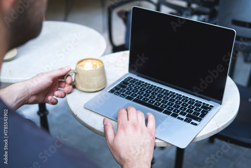 Male hands working with laptop and coffee cup on round table in outdoor cafe.