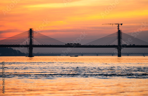 A boat sails on the river Brahmaputra during  sunset, with the silhouette of a cable-stayed bridge and hills in the background
