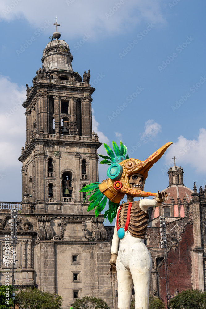 Fototapeta premium A skeleton dressed in a costume with a bird head and a skeleton's face is standing in front of a lar. Mexican Day of the Dead tradition, offerings, food, colors, decorations, and papel picado, La Catr