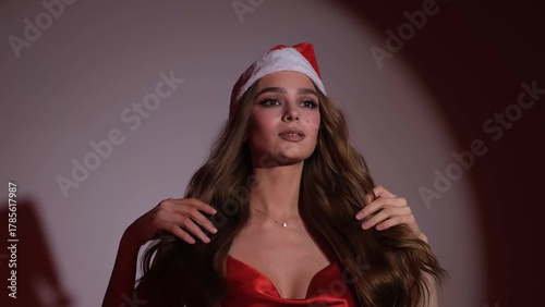 A sexy happy woman with long hair in a New Year's hat and a red dress poses on a neutral background at a New Year's party-style photo shoot