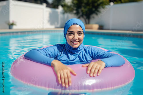 Muslim woman in burkini swimwear enjoying floating in swimming pool, using inflatable pink swim ring.