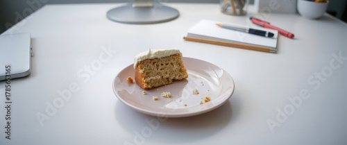 A slice of cake on a pink plate sits on a white table. Nearby are a notebook, pen, and a desk lamp, creating a cozy workspace atmosphere.