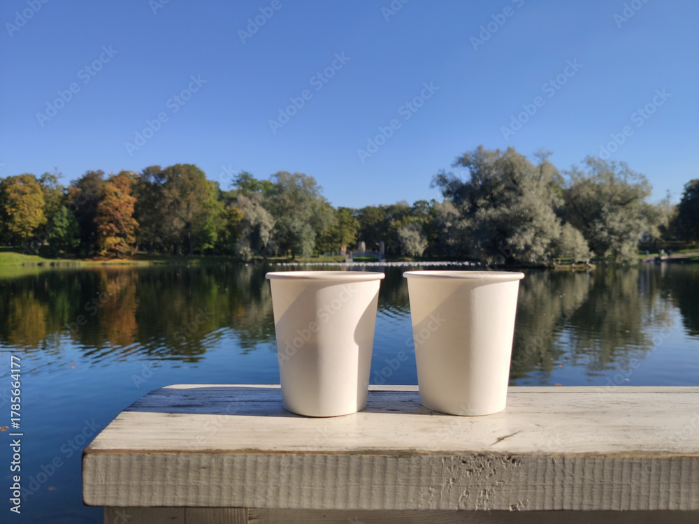 Obraz premium Two white paper cups are placed on a wooden table, with a view of a lake and a park. Picturesque nature. A cozy outdoor recreation. Gatchina Palace Park. Russia.