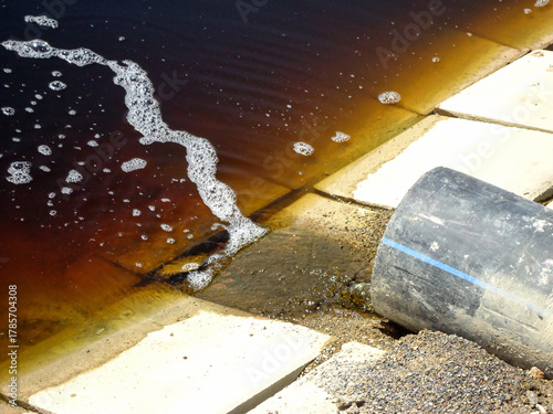 Detailed photo of heavily polluted water landfill leachate draining from pipe into evaporation pond with tiled edges, showing waste management, environmental contamination, industrial water treatment