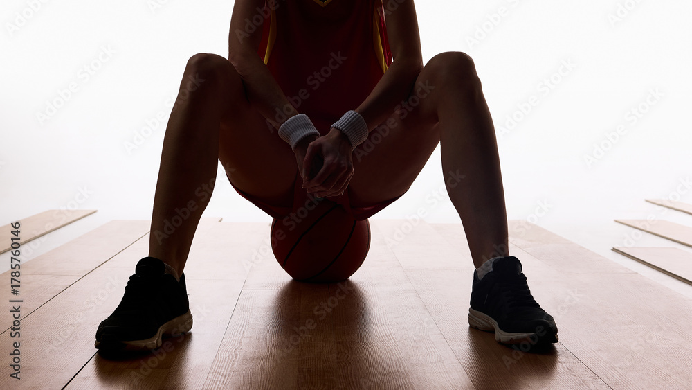 Naklejka premium Female basketball player sitting with ball between knees in focus moment. Concept of concentration, discipline, training motivation, and sports advertising visuals.