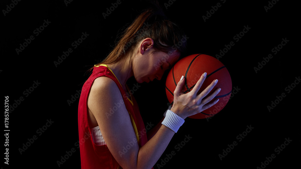 Obraz premium Female basketball player resting forehead on ball in emotional reflection. Concept of inner strength, healing, and balance for sports health and motivation media.