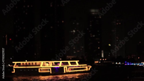 Slow motion, abstract view of a brightly illuminated tour boat cruising on the water at night. Defocused city skyscrapers form a beautiful bokeh background