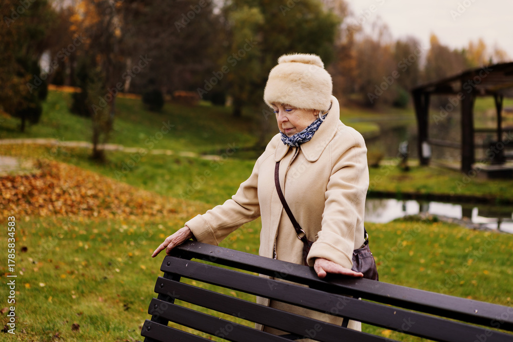 Fototapeta premium Sad elderly woman standing alone in autumn park, looking down with thoughtful expression. Depressed senior female outdoors surrounded by golden trees and fallen leaves, symbol of loneliness and aging