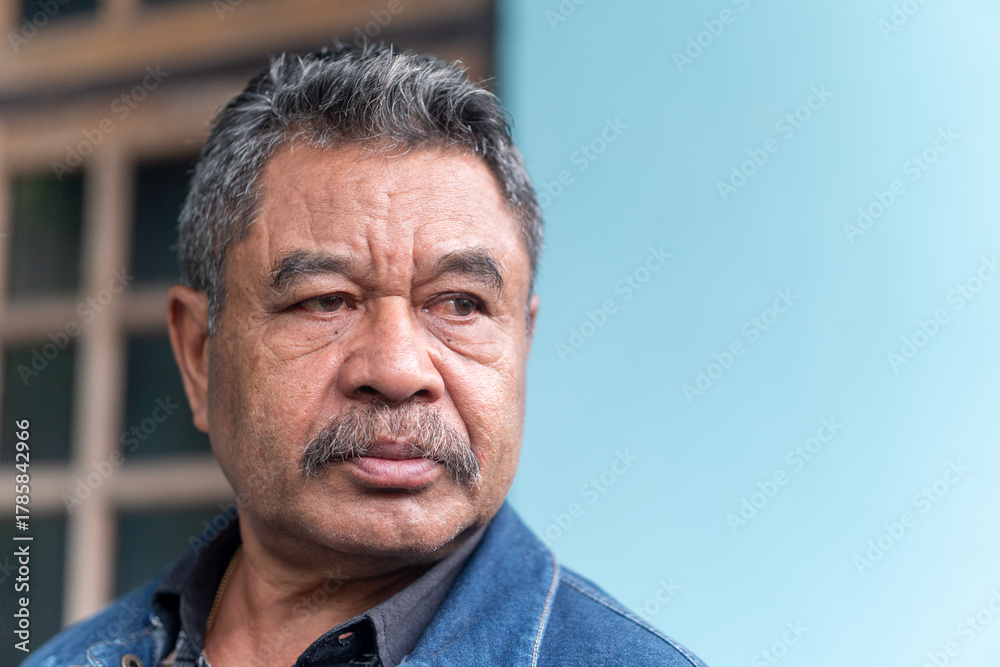 Obraz premium A candid outdoor portrait of a thoughtful senior Asian man. He has a pensive expression, wearing a denim jacket.
