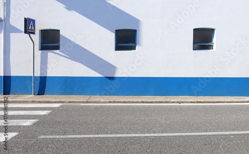 Fototapeta Naklejka Na Ścianę i Meble -  Blue and white plaster wall with three small windows. Sidewalk and street in front.Pedestrian crossing sign and crosswalk on the left. Background for copy space