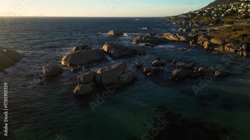 Aerial view of large boulders sitting in the ocean water along a rocky coastline with buildings on the hilltop, Cape Town, Western Cape, South Africa.