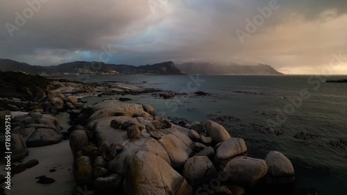 Aerial view of massive boulders scattered along the tranquil coastline with a backdrop of mountains under dramatic skies, Cape Town, Western Cape, South Africa.