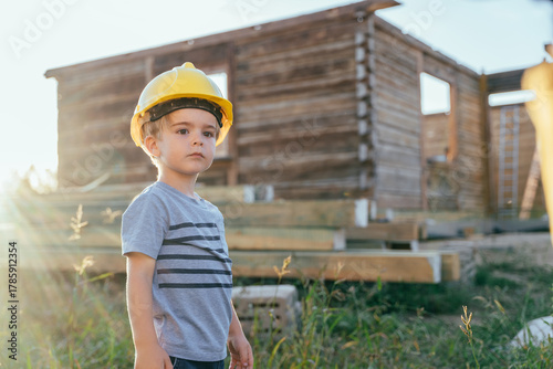 Little boy wearing construction helmet standing near wooden house in sunlight