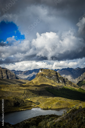Cajas national park. Mountain landscape under dramatic cloudy sky with sunlight breaking through. Scenic highland view with hills, rocky peaks, and calm lake in natural light