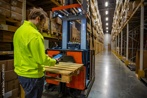 Warehouse employee using a tablet to operate a pallet stacker in a large storage facility. The worker in a high-visibility jacket checks inventory among tall shelves filled with cardboard boxes.