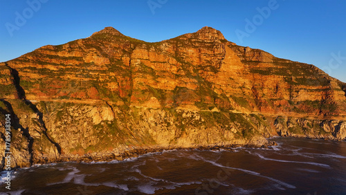 Aerial view of majestic mountains meet the restless ocean in a symphony of golden light and rugged textures, Cape Town, Western Cape, South Africa.