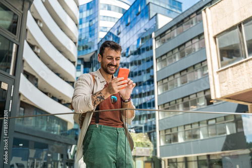 Young traveler laughing while taking a selfie in a modern business district cheerful millennial man with backpack capturing urban architecture and city vibes on smartphone during a sunny day