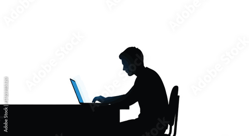 Silhouette of a young man focused on typing on a laptop computer at a desk with a bright screen