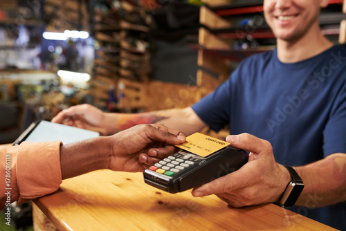 Young man holding payment terminal while Black young man making contactless payment with credit card at counter in retail store, both smiling during transaction